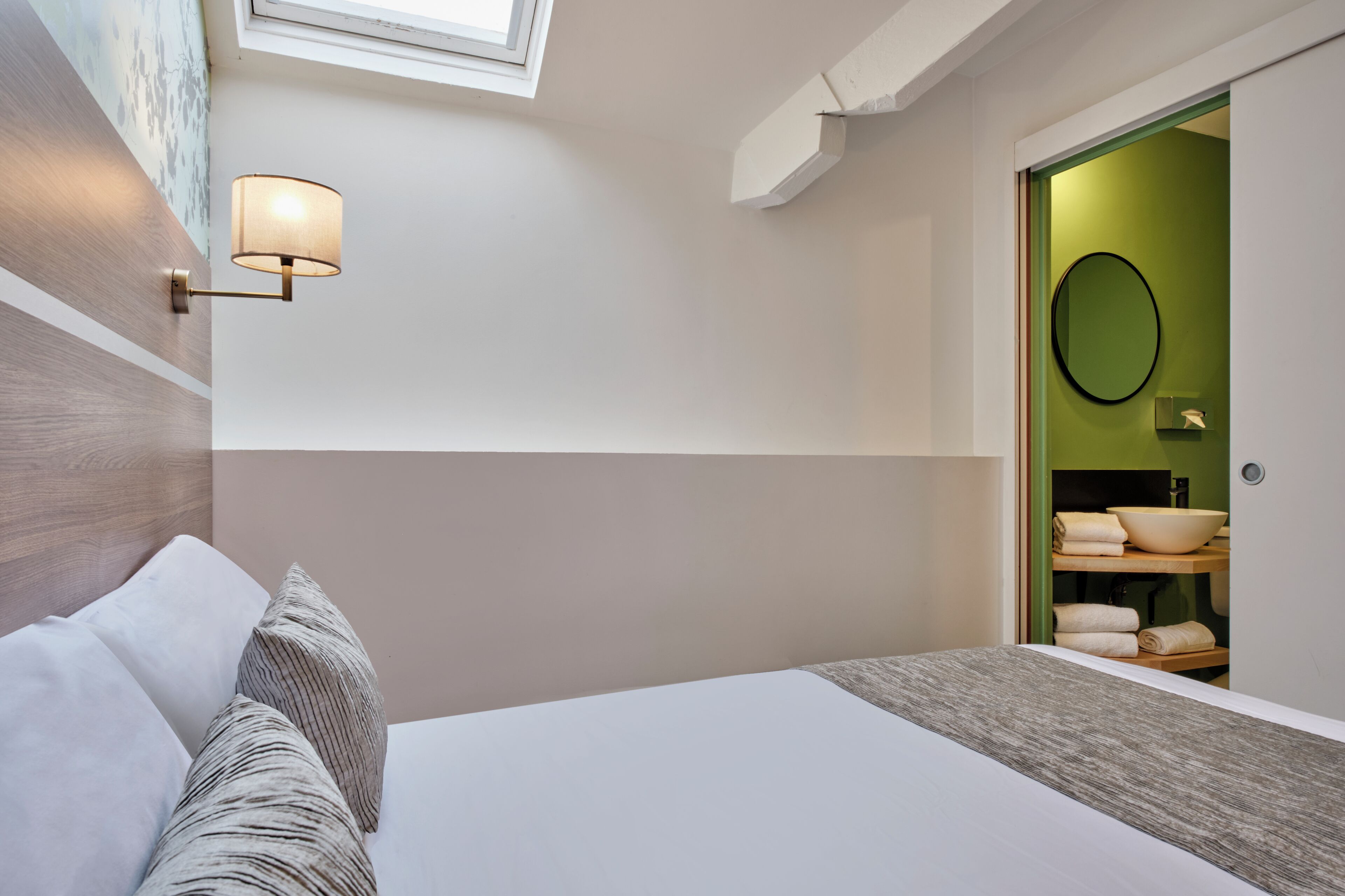 Un dormitorio moderno con tragaluz, una lámpara de noche, una cama con almohadas y vistas a un baño con pared verde y espejo redondo.