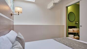 Un dormitorio moderno con tragaluz, una lámpara de noche, una cama con almohadas y vistas a un baño con pared verde y espejo redondo.