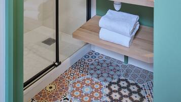 A bathroom with a glass shower enclosure, a white sink with a wooden countertop, a round mirror, and a patterned tile floor.