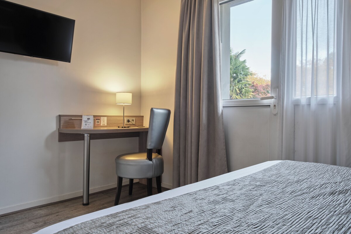 A hotel room with a bed, a desk with a chair, a flat-screen TV, a lamp, and a window with curtains.
