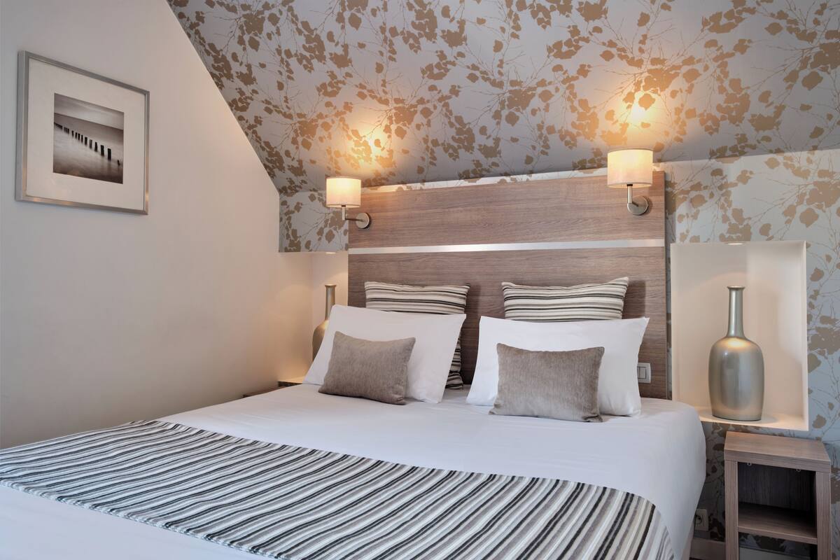 A hotel room with a bed, two wall-mounted lamps, a framed picture, and a decorative vase on a nightstand.