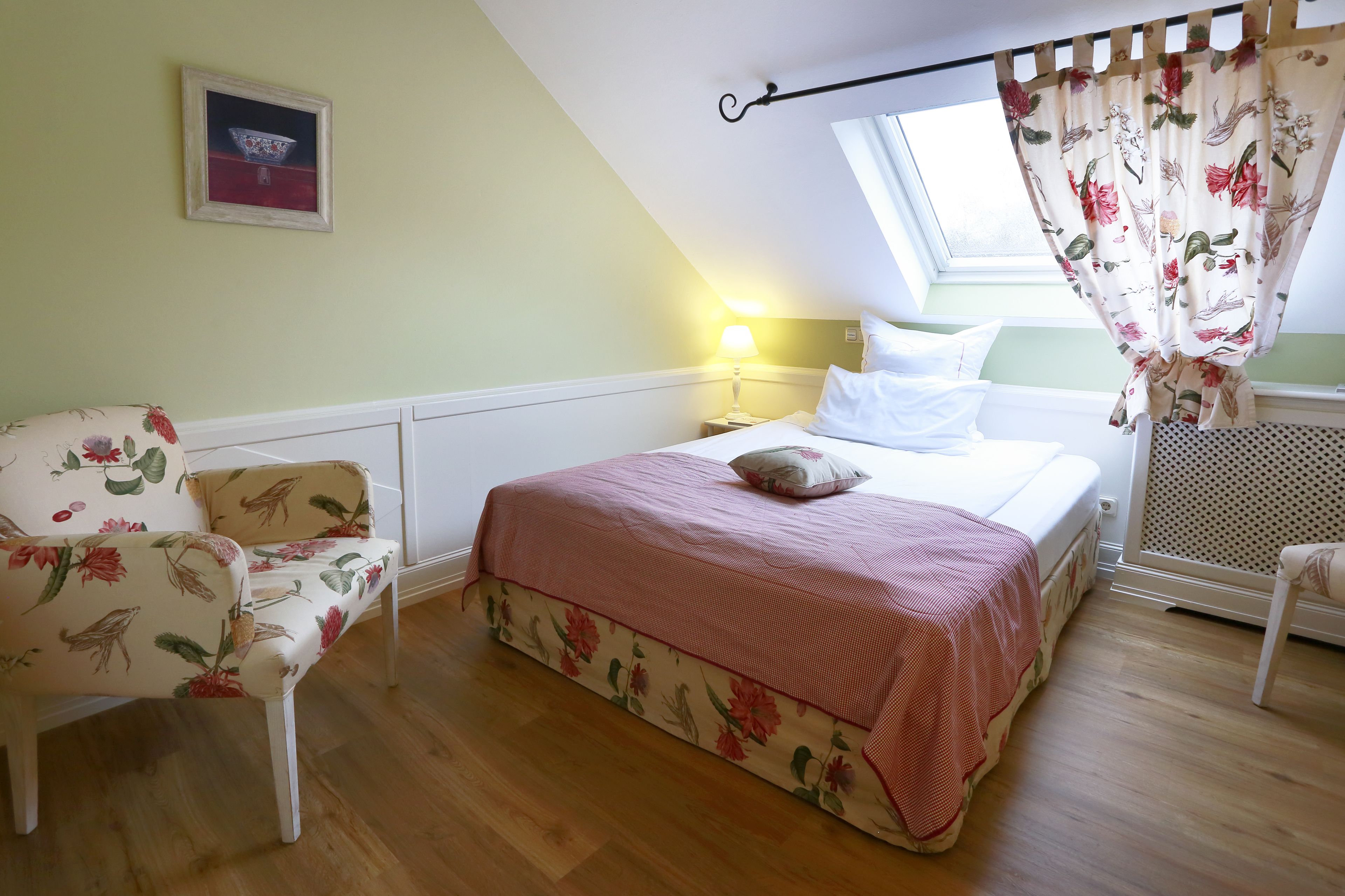 Une chambre à coucher avec un lit, une chaise, une table de chevet, une lampe, une fenêtre avec des rideaux et un tableau au mur.