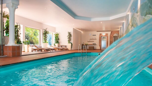 Una piscina interior con una cascada, rodeada de sillones reclinables y plantas en macetas.