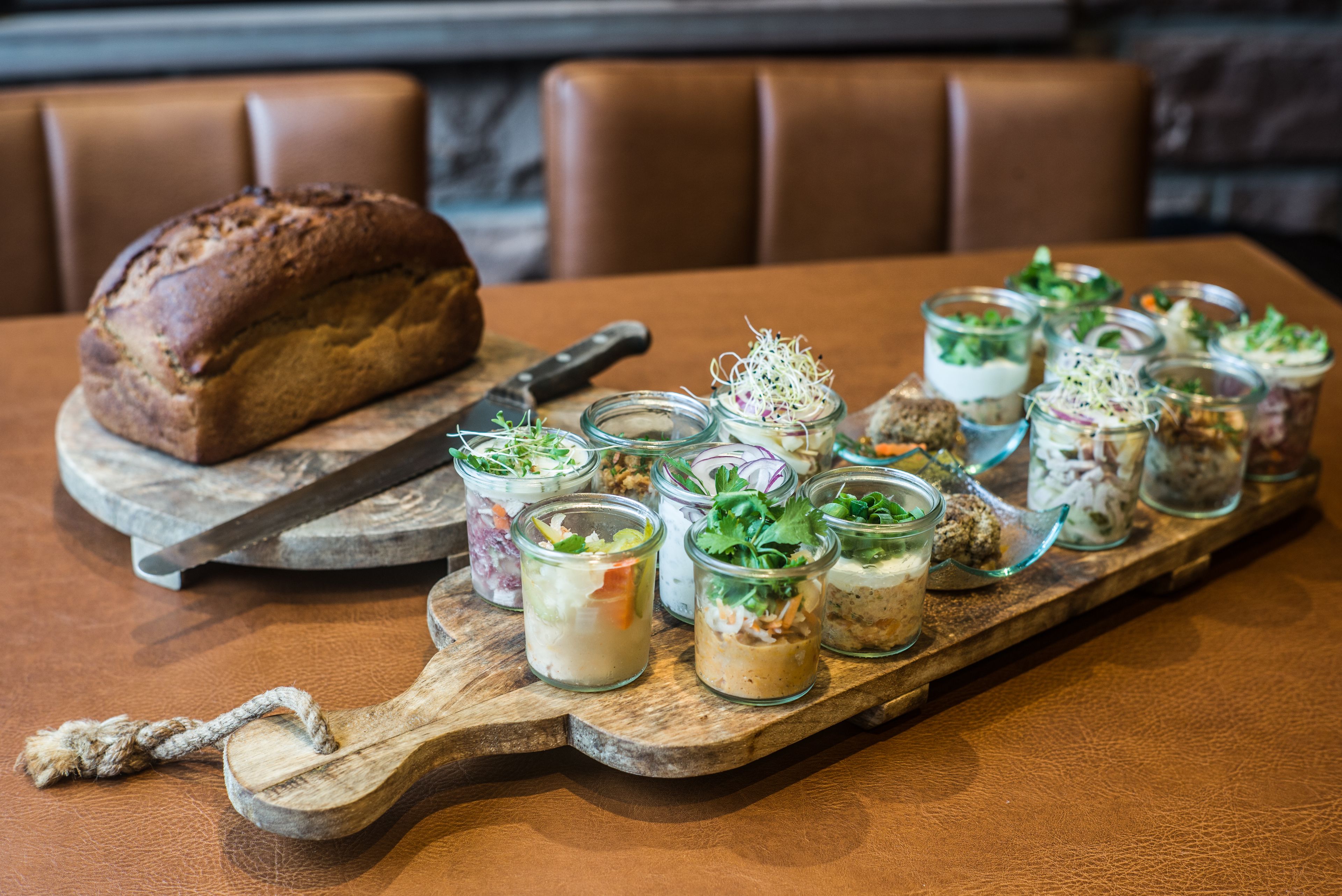 A wooden serving board with various food items and a loaf of bread on a table.