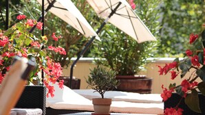 Un patio con dos sombrillas color beige, una planta pequeña en maceta y flores rojas.
