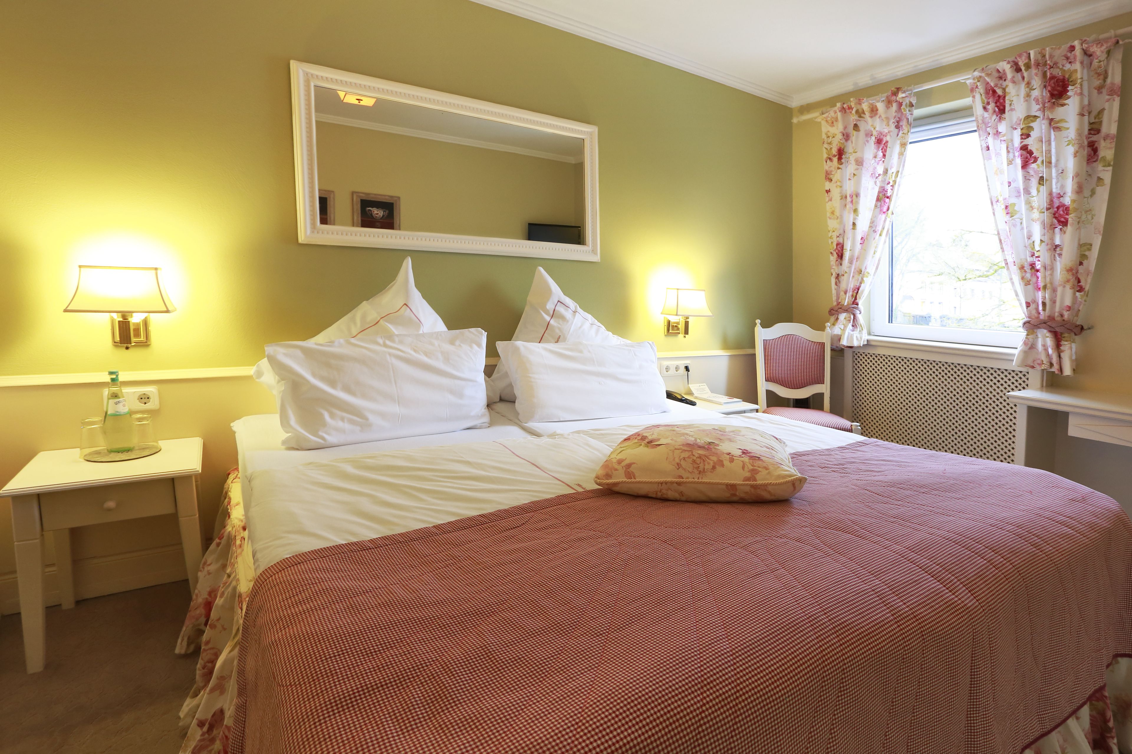 A neatly arranged hotel room with a large bed, two bedside tables with lamps, a chair, a window with floral curtains, and a mirror on the wall.