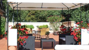 Un patio al aire libre con una zona de estar cubierta, que incluye una mesa y sillas, rodeado de macetas con plantas y flores.