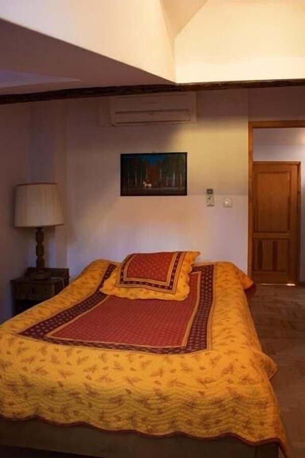 Un dormitorio con una cama cubierta por una colcha estampada, una mesita de noche con una lámpara, un cuadro en la pared y una puerta de madera.