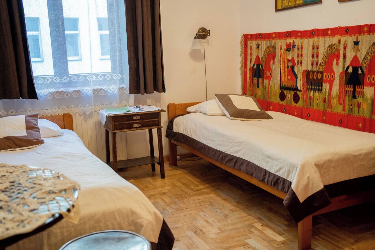 A room with two single beds, a small wooden table, a lamp, and a colorful tapestry on the wall.