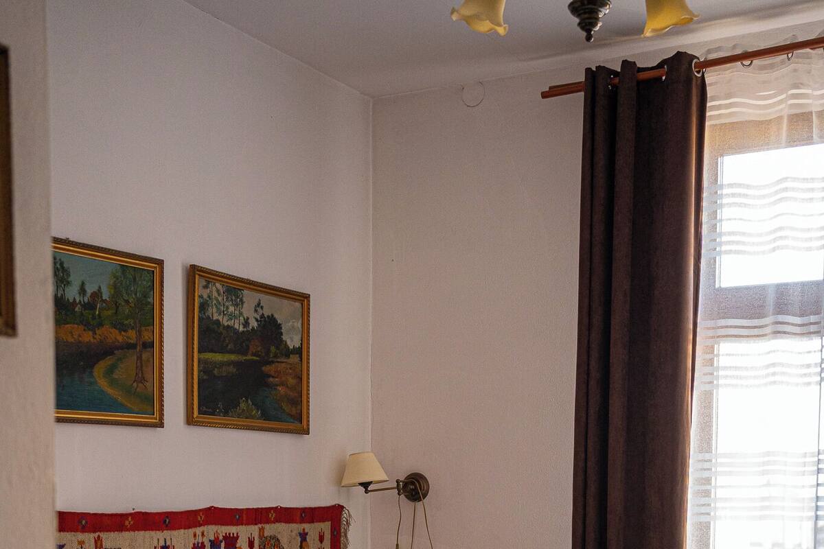 A bedroom with a bed, wooden floor, two paintings on the wall, a lamp, and a window with curtains.