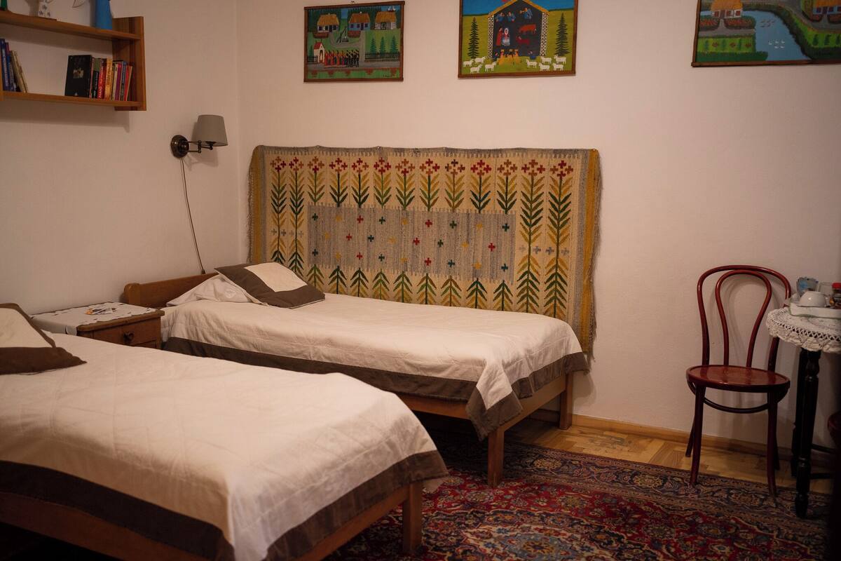 A room with two beds, a small table with a chair, a lamp, and a bookshelf.