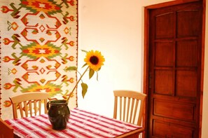 A table with a red and white checkered tablecloth, a dark ceramic pitcher, and a single sunflower. Two wooden chairs are positioned around the table. A wooden door is visible in the background. - Kolory Guest House (Kraków)