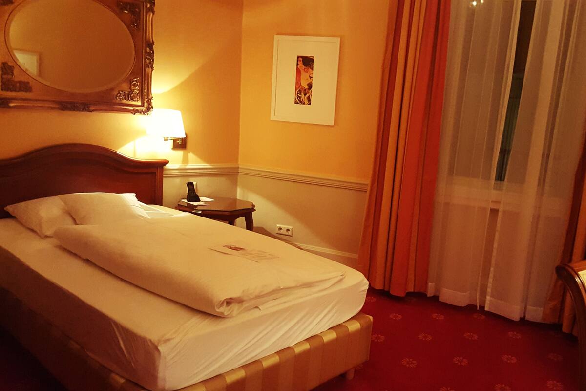 A hotel room with a bed, a bedside table with a lamp, a mirror, a framed picture on the wall, and a window with curtains.
