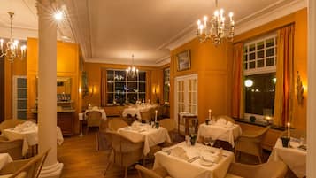 Comedor con mesas dispuestas para varios comensales, candelabros y vistas al exterior a través de amplios ventanales.
