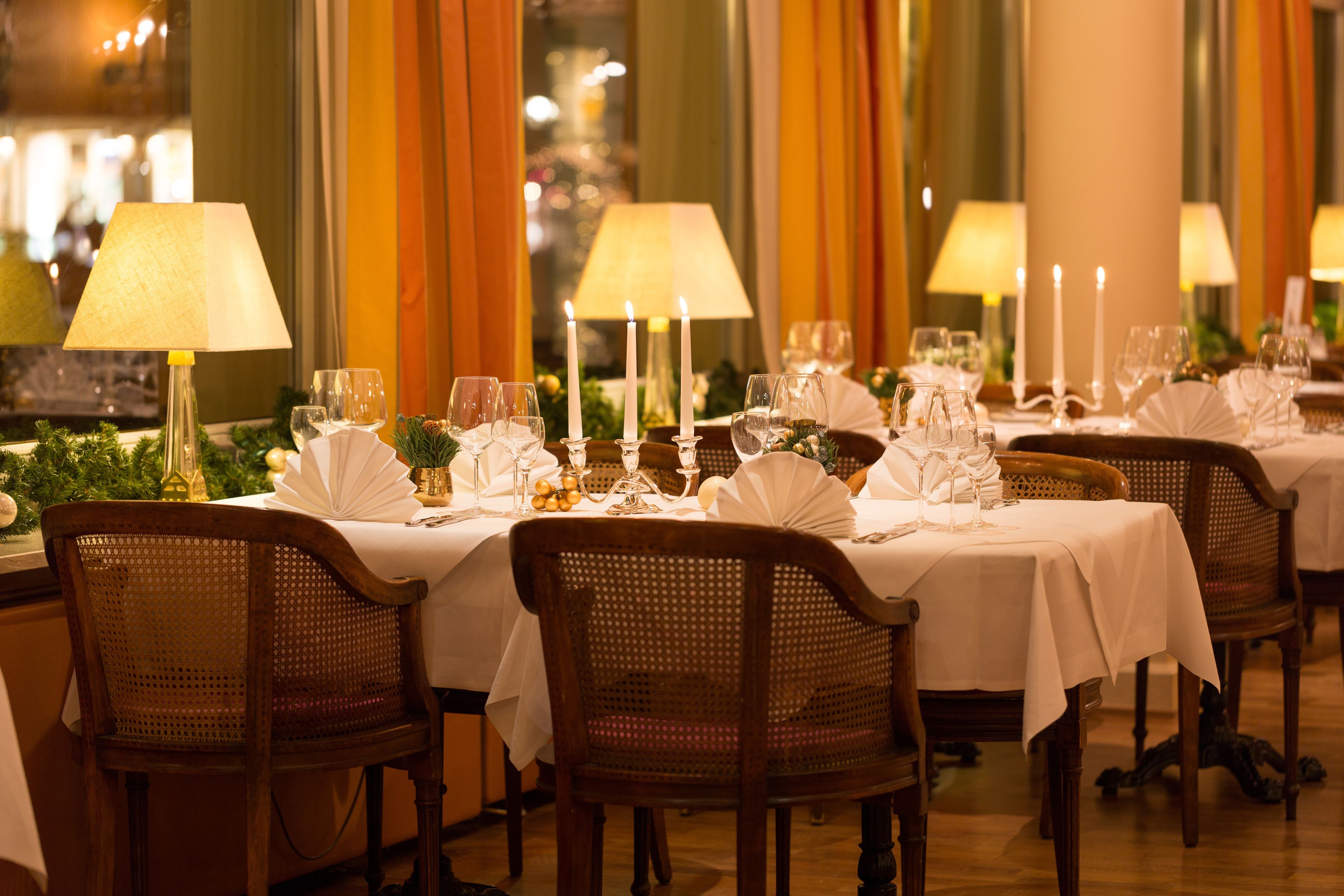 Un restaurant où de nombreuses tables sont dressées pour le service, chacune étant recouverte d’une nappe blanche, agrémentée de bougies et de verres.