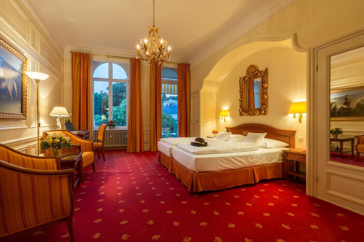 A hotel room with a large bed, a desk with a lamp, a chair, a window with curtains, and a chandelier.