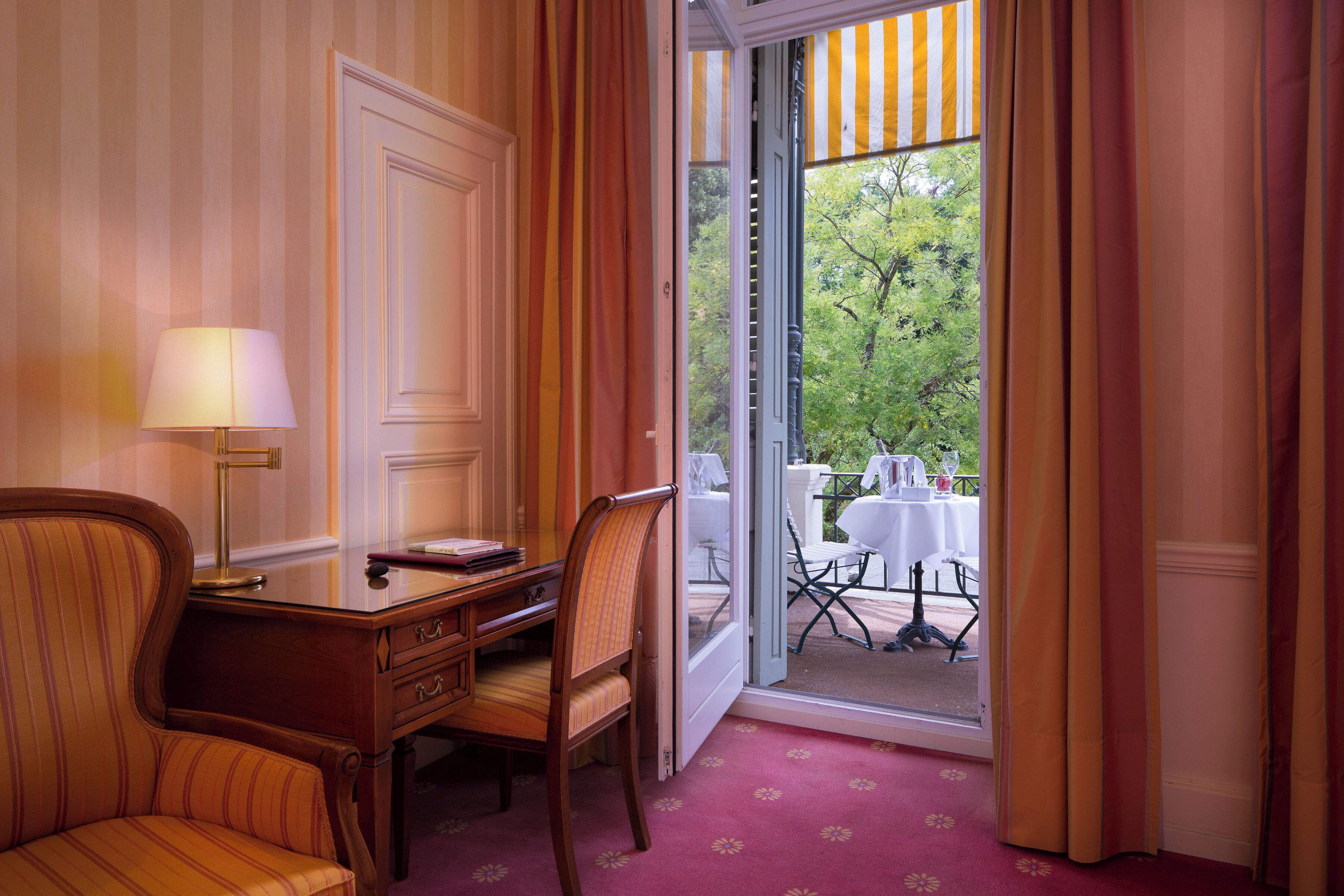 A hotel room with a desk, two chairs, a lamp, and a view of an outdoor dining area.