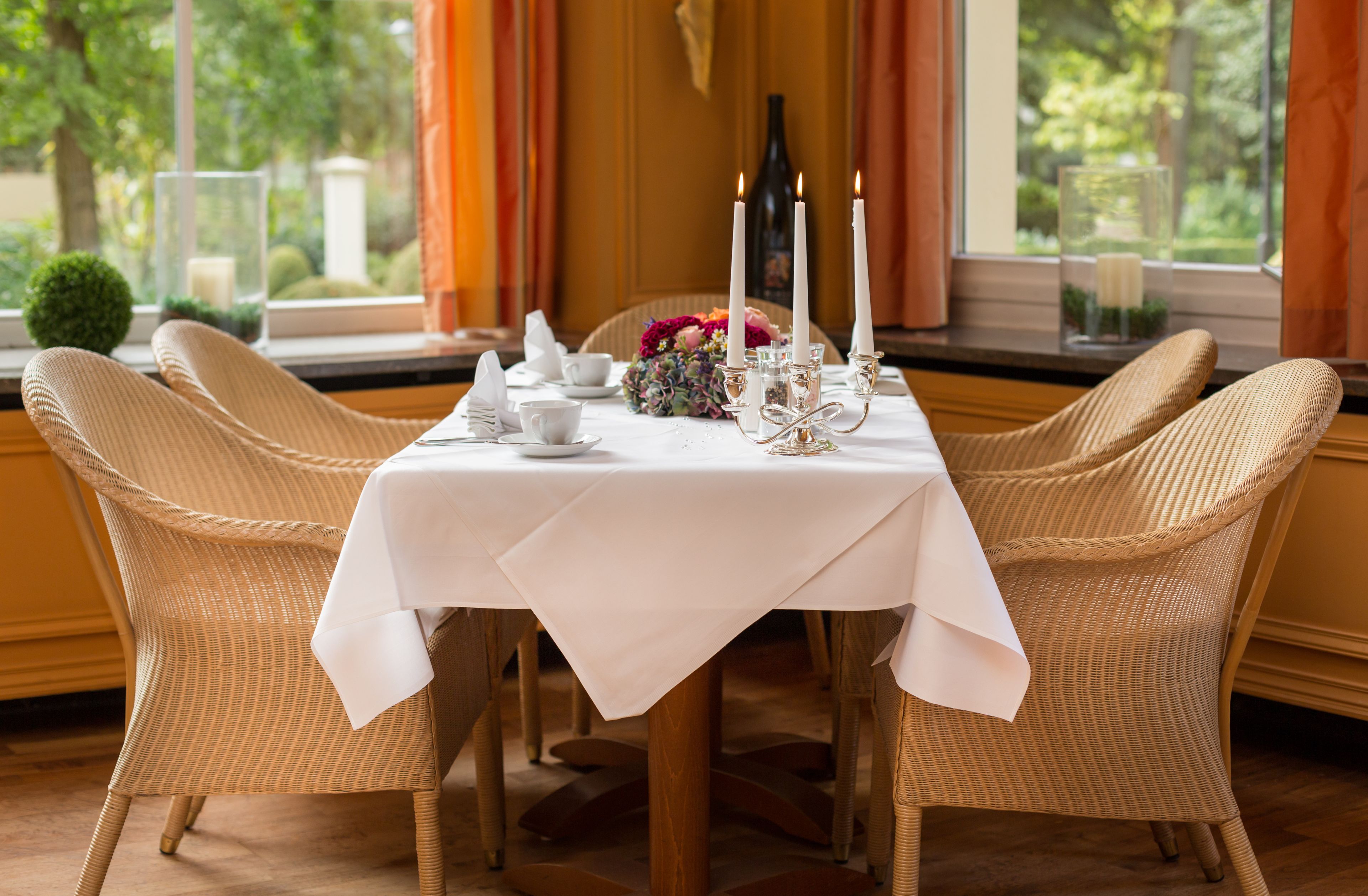 A dining table set with white tablecloth, wicker chairs, candles, and a vase of flowers.