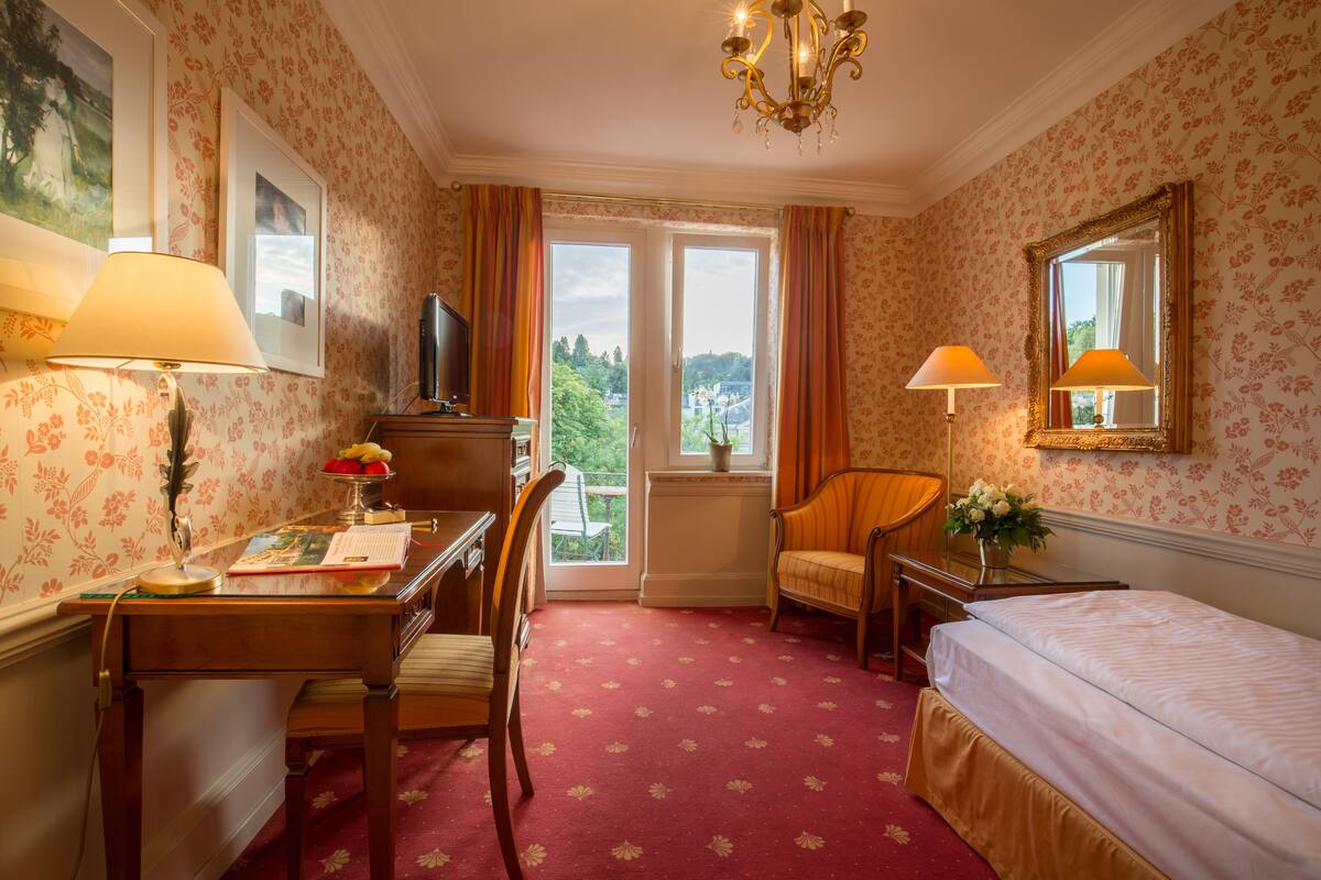 A hotel room with a desk, chair, bed, and a view of the outdoors through a window.