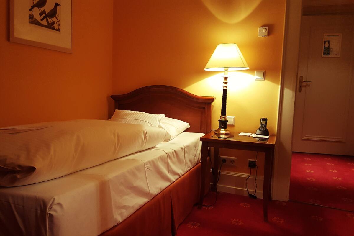 A hotel room with a bed, a bedside table with a lamp, a telephone, and a door.