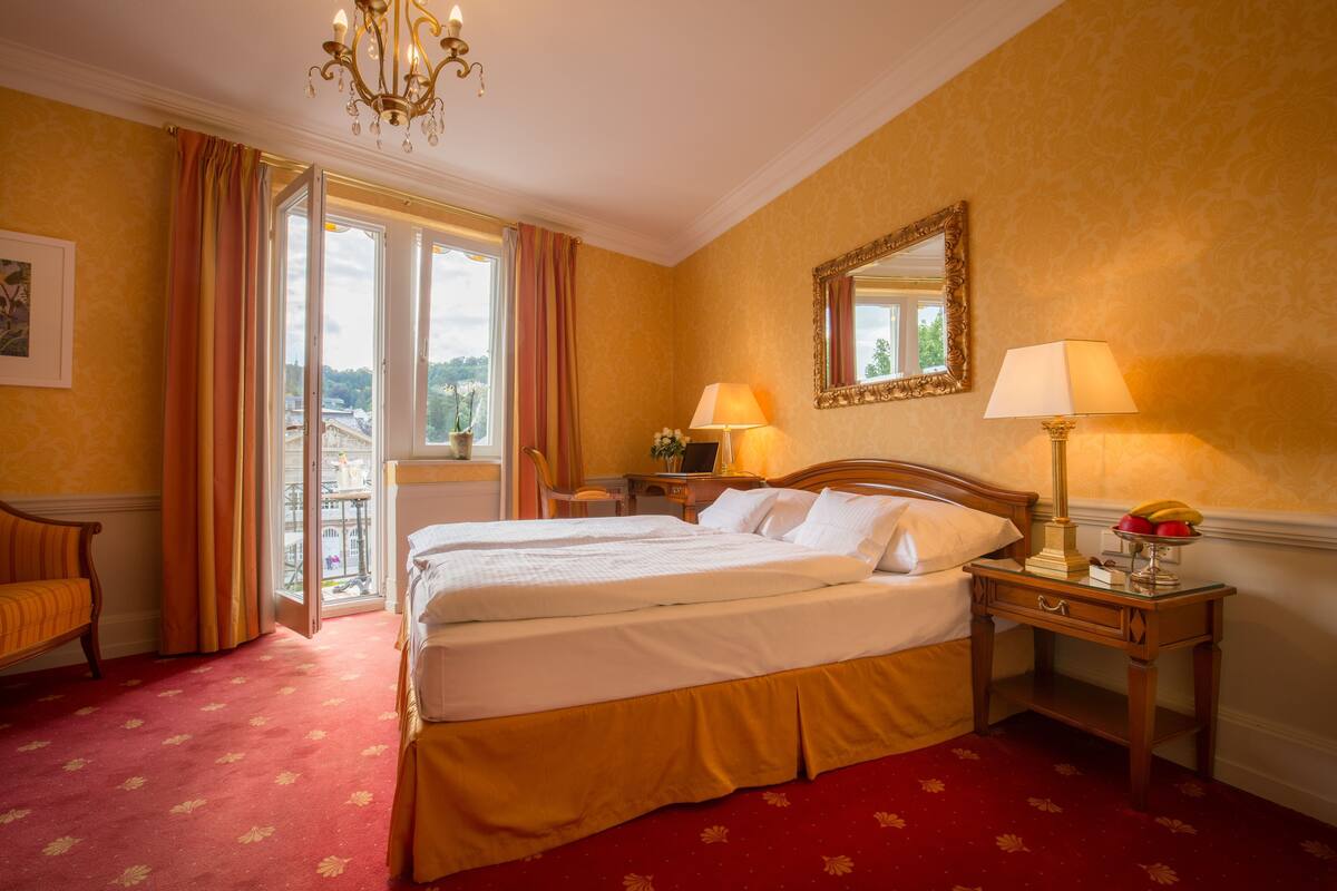 A hotel room with a bed, two bedside tables, a chandelier, a window with curtains, and a mirror on the wall.