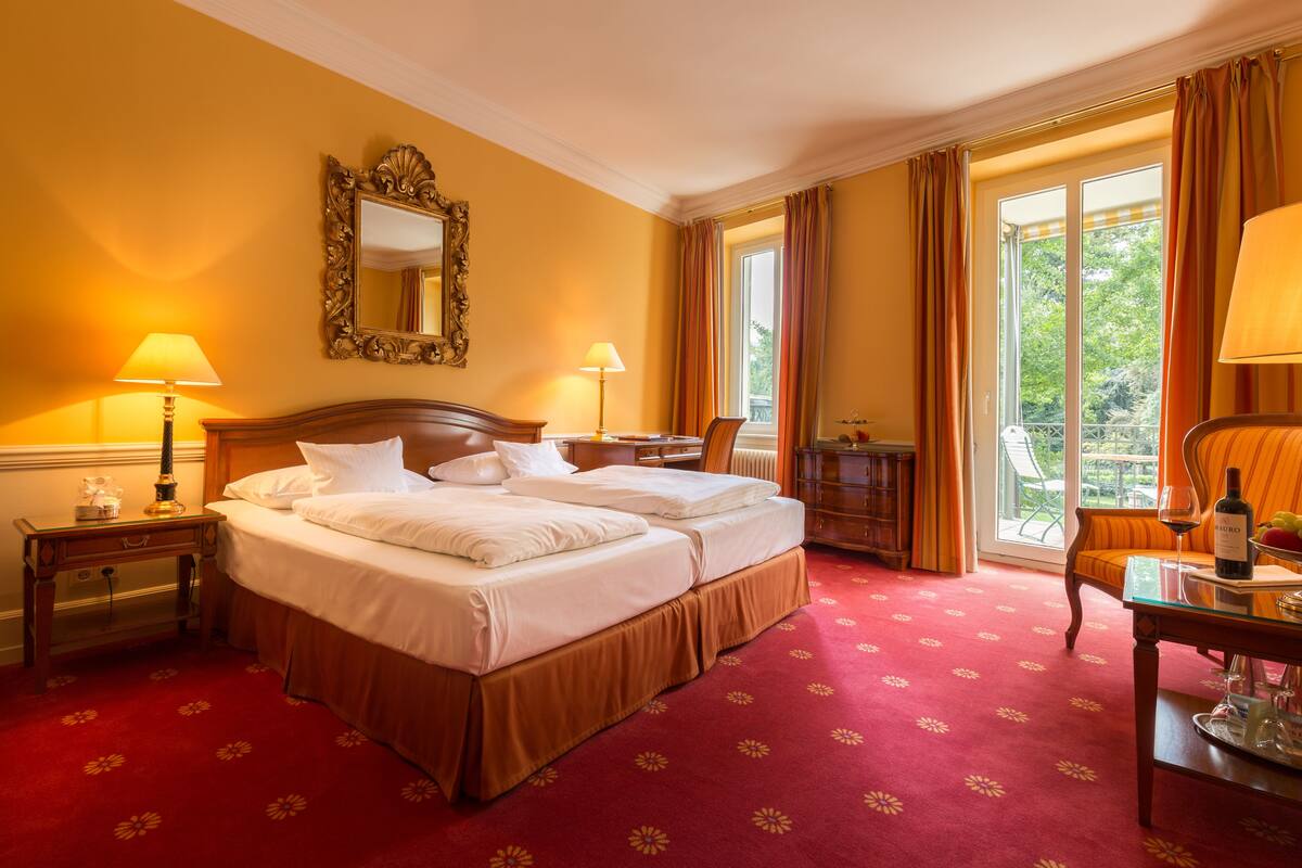 A hotel room with a large bed, two bedside tables with lamps, a chair, a small table with a wine bottle, and a view of greenery through the window.