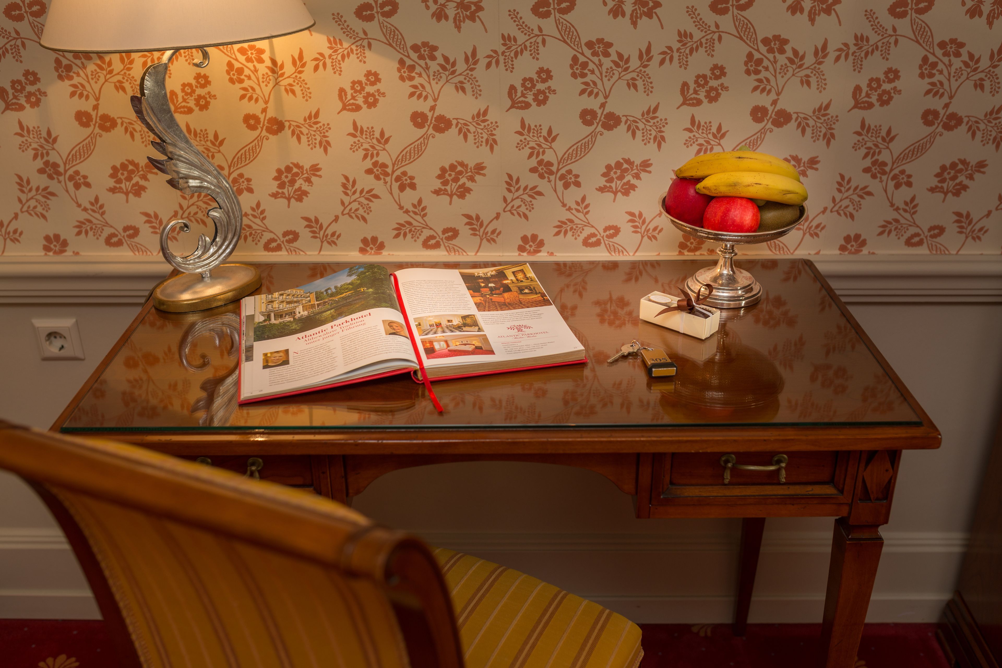 A wooden table with a glass top, a lamp, an open book, a fruit bowl, and a chair.
