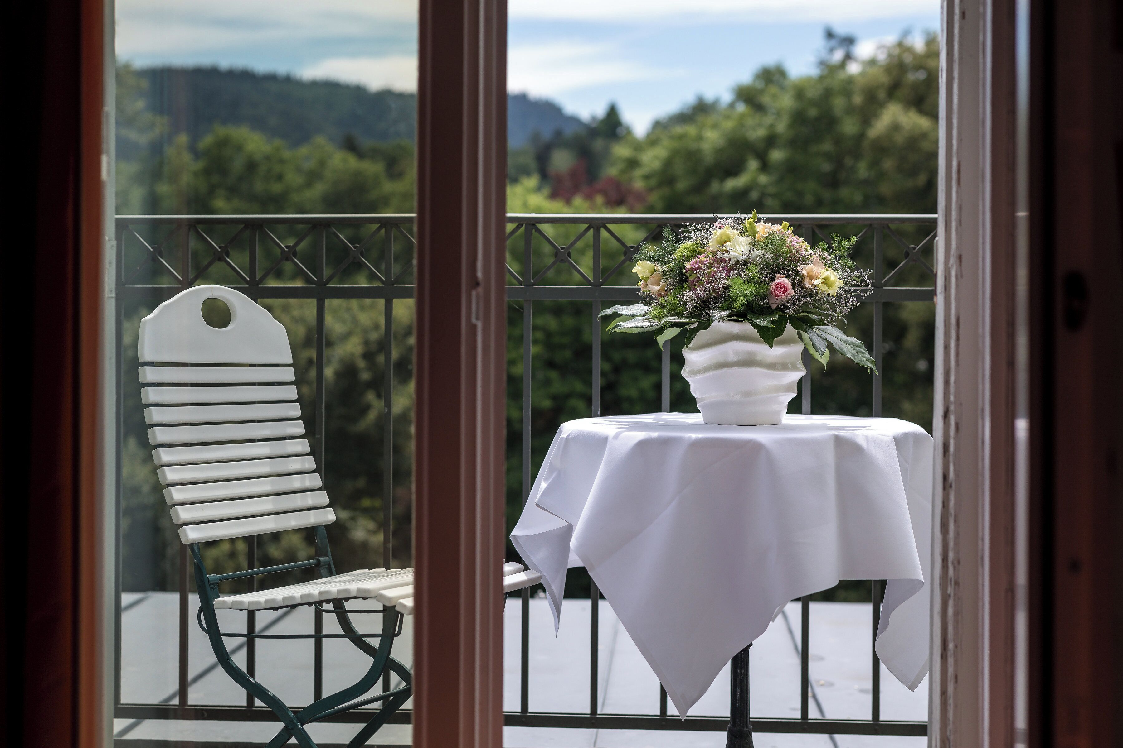 A balcony with a white table and chair, a vase of flowers, and a view of trees and hills.