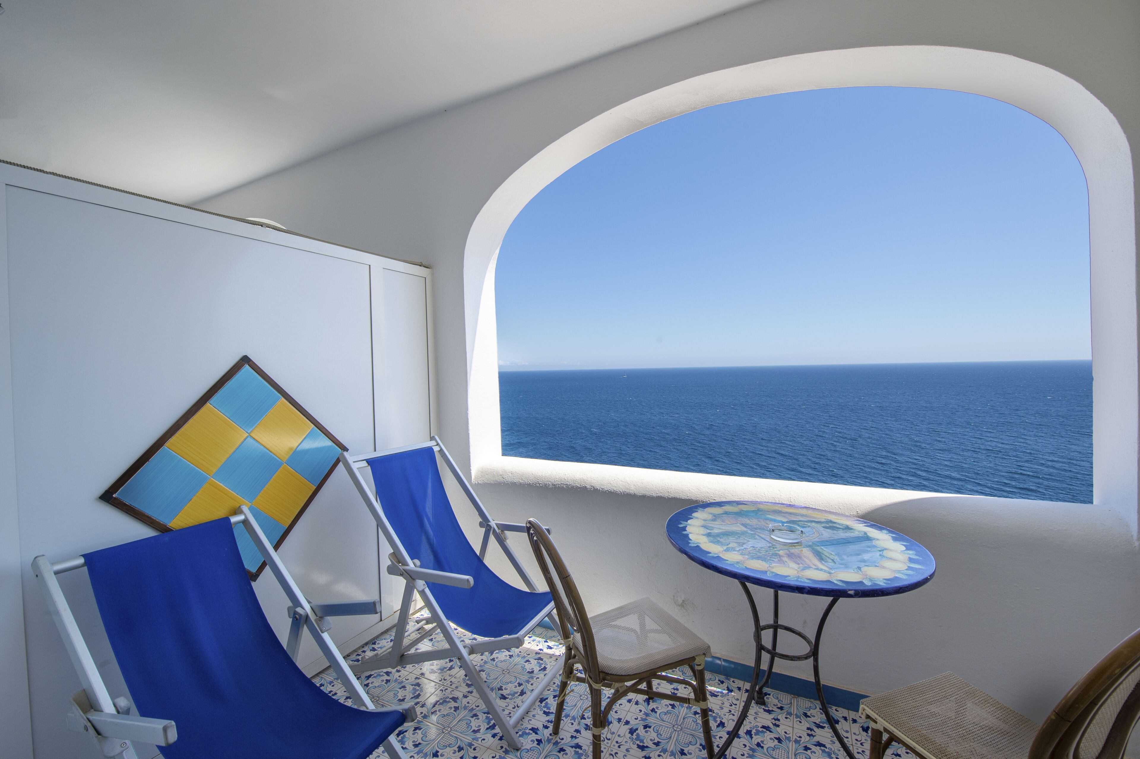 A balcony with a view of the sea, blue chairs, a small table, and a decorative wall piece.