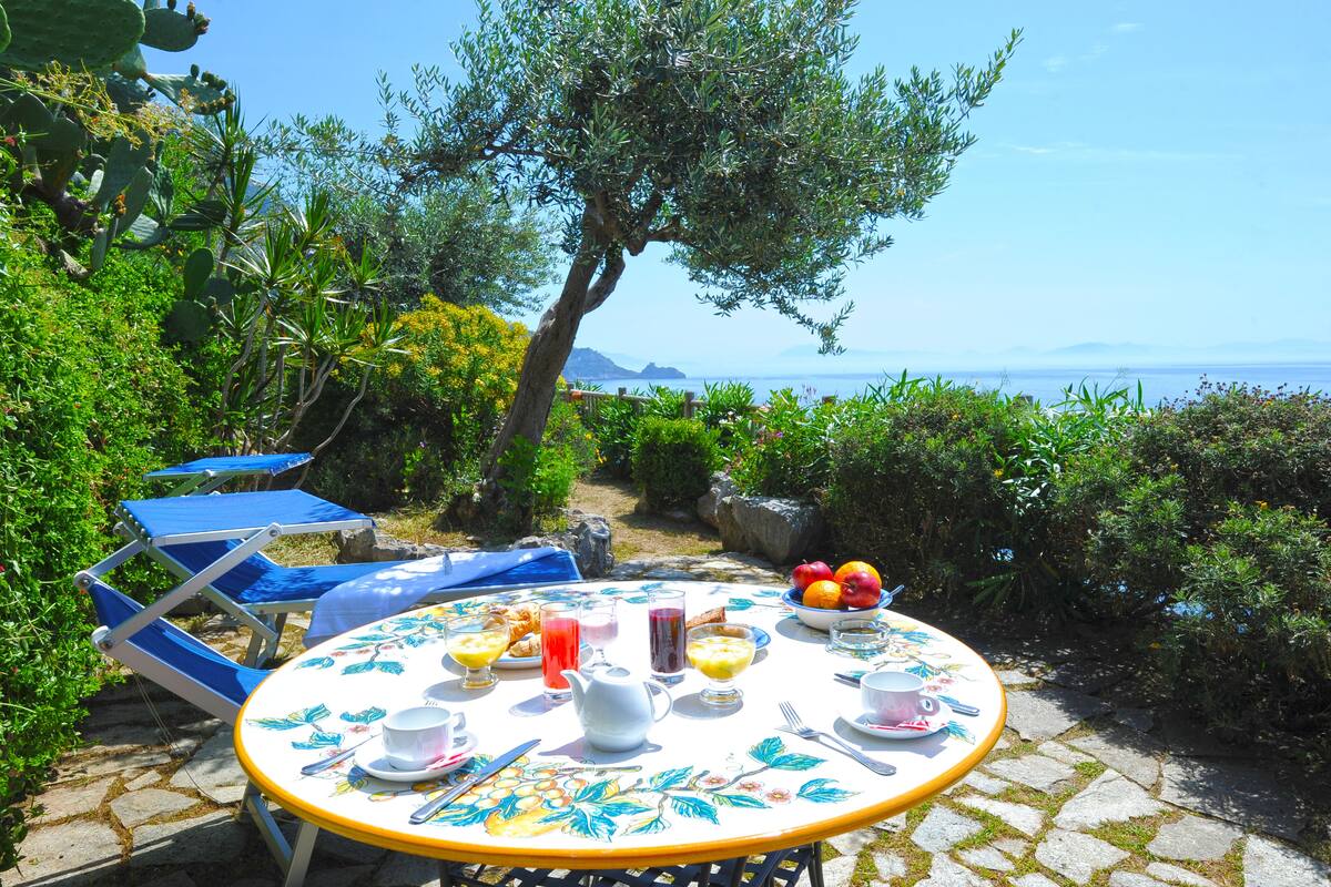 Una mesa redonda con mantel floral, rodeada de sillas azules, ubicada al aire libre con vista al mar y montañas al fondo.