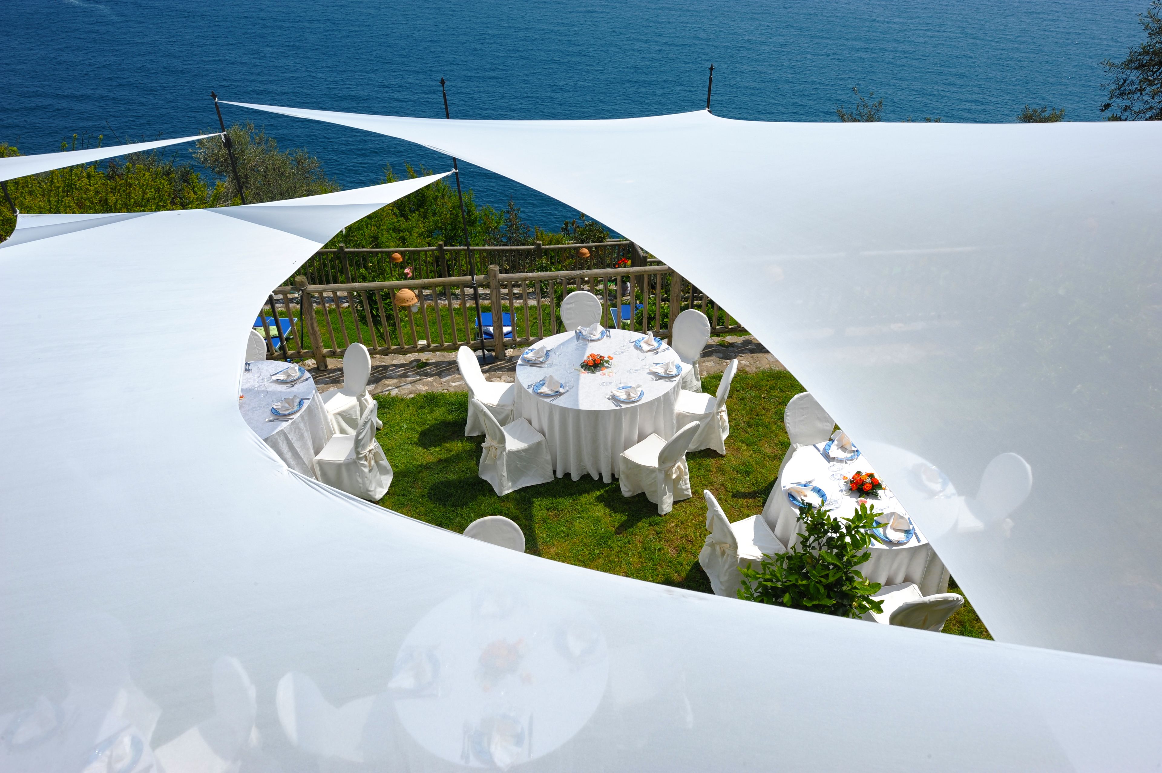 Uma área de refeições ao ar livre com mesas redondas e cadeiras brancas, montada em um terraço gramado com vista para o mar.