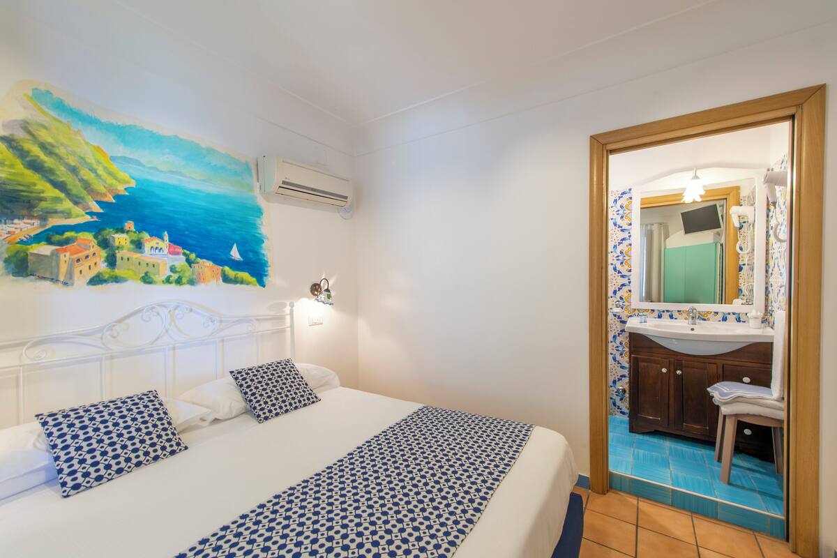 Habitación de hotel con una cama, un cuadro de una escena costera, un baño con lavamanos y espejo, y suelo de baldosas.
