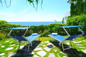 Two sun loungers with blue cushions and a pair of flip-flops on a stone pathway leading to a view of the sea.