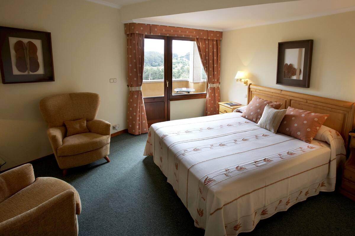 Una habitación de hotel con una cama, dos sillas, una mesita de noche con lámpara y un ventanal con cortinas.