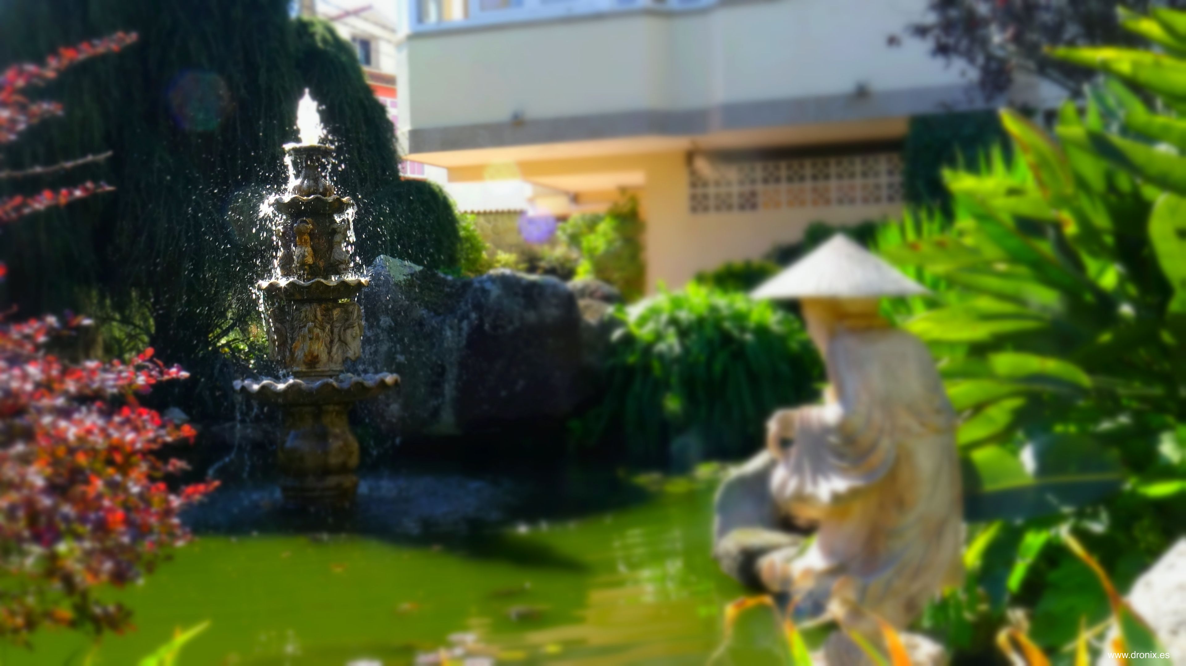 A fountain with water spouting from multiple tiers, surrounded by greenery and a statue.