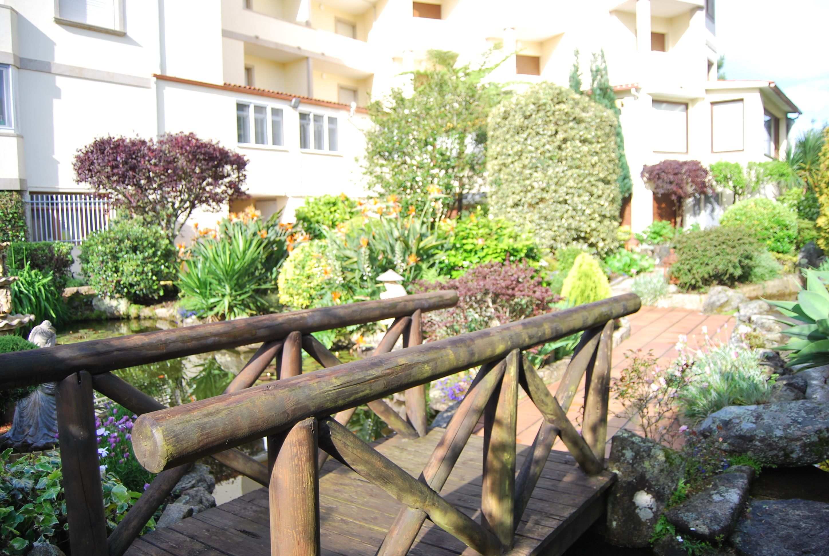 Un puente de madera en un jardín con diversas plantas y un edificio al fondo.