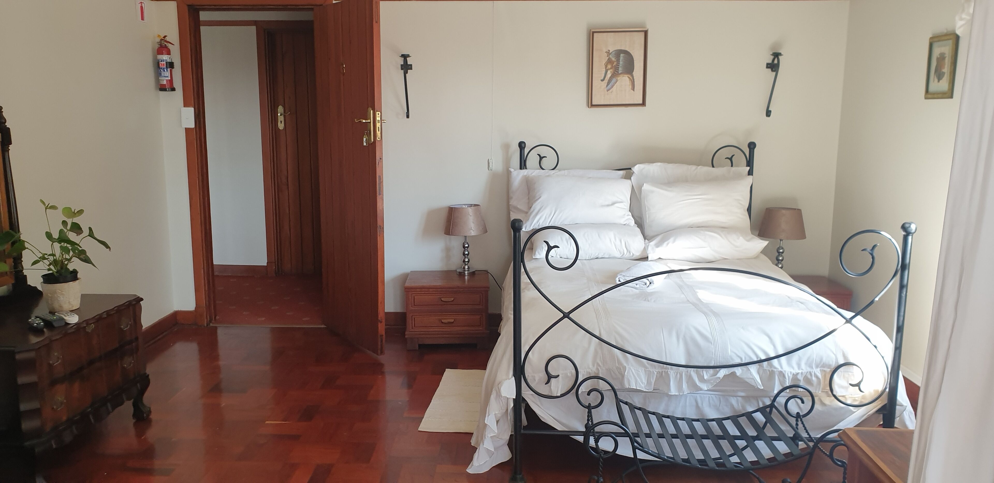 Un dormitorio con estructura de cama metálica, ropa de cama blanca, una mesita de noche con lámparas, un aparador de madera y una puerta que da a otra habitación.