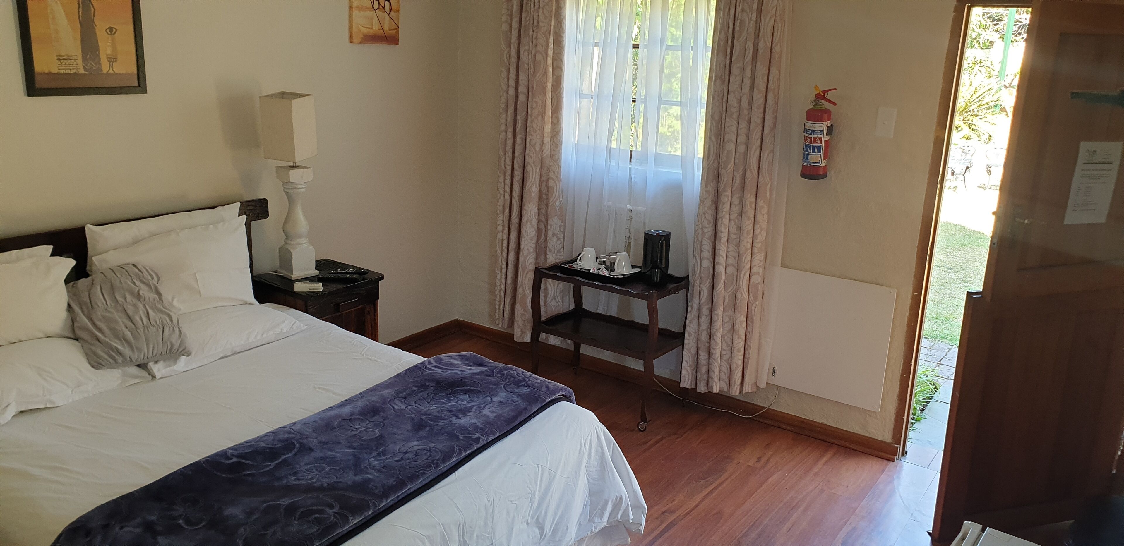Une chambre d’hôtel avec un lit, une table de chevet avec une lampe, une petite table avec des tasses et une bouilloire, et un extincteur fixé au mur.