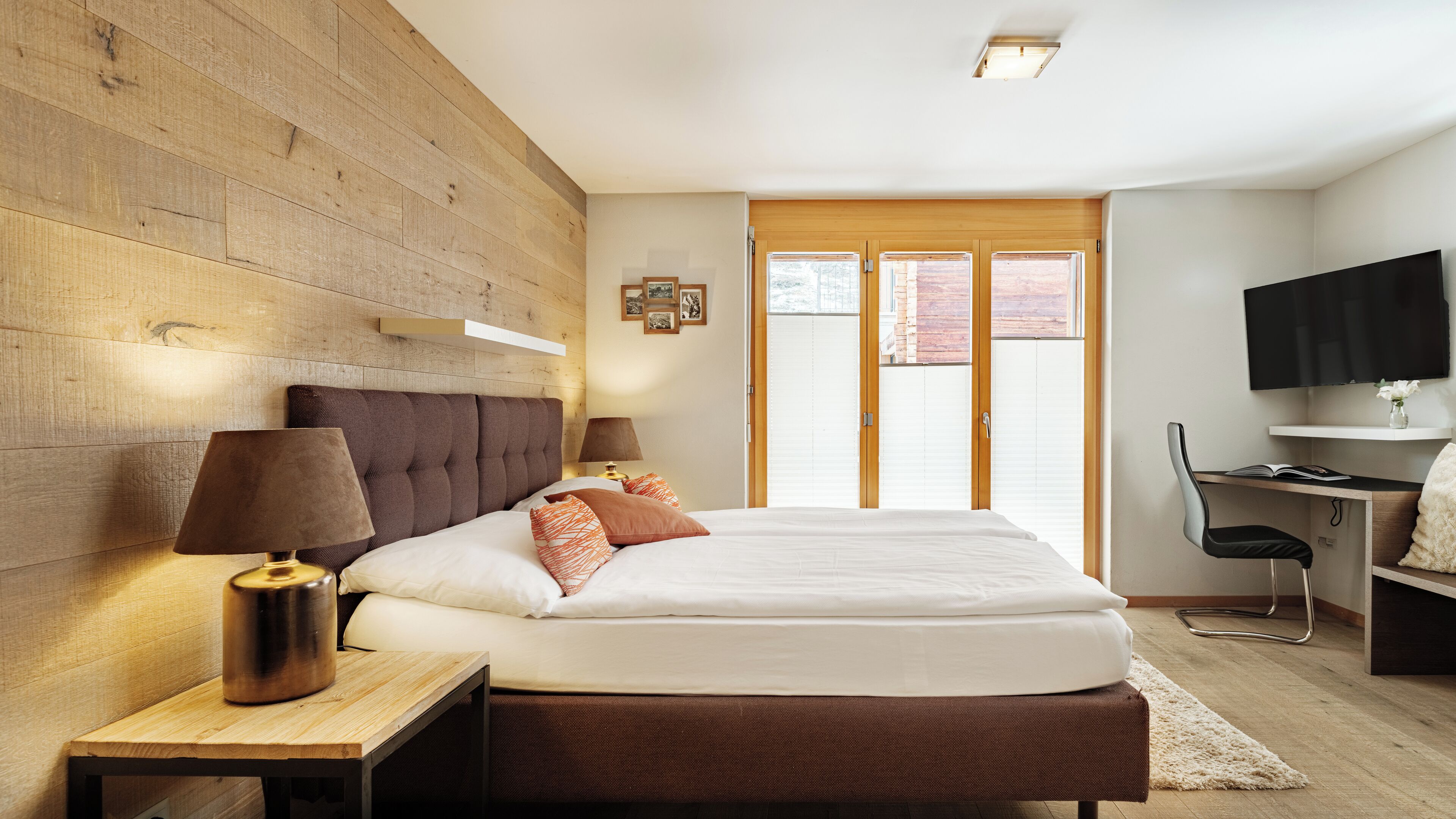 A modern bedroom with a large bed, wooden headboard, bedside lamps, a desk with a chair, and a TV.