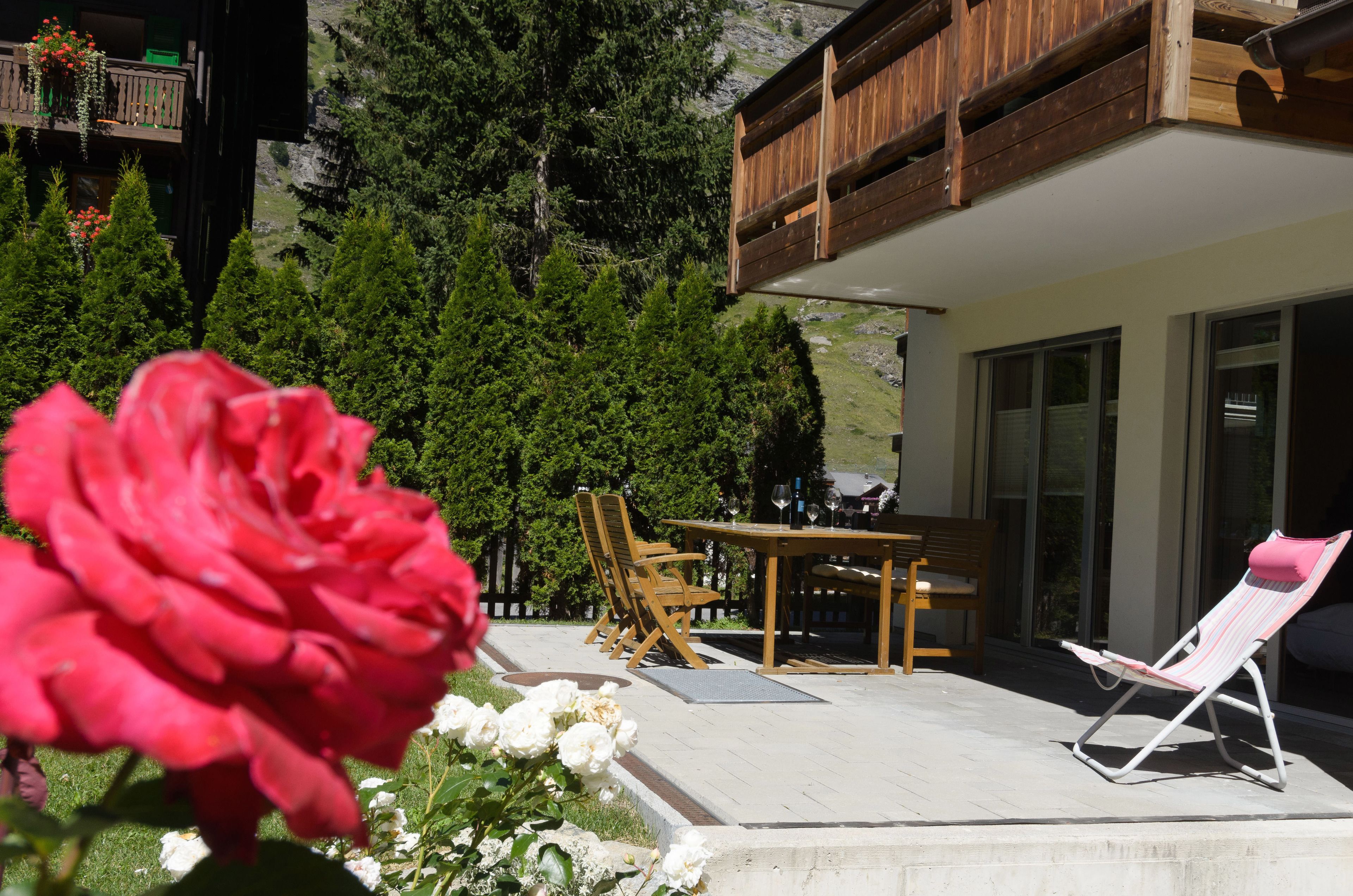 A patio with a table and chairs, a lounge chair, and a wooden deck overlooking a forested area.