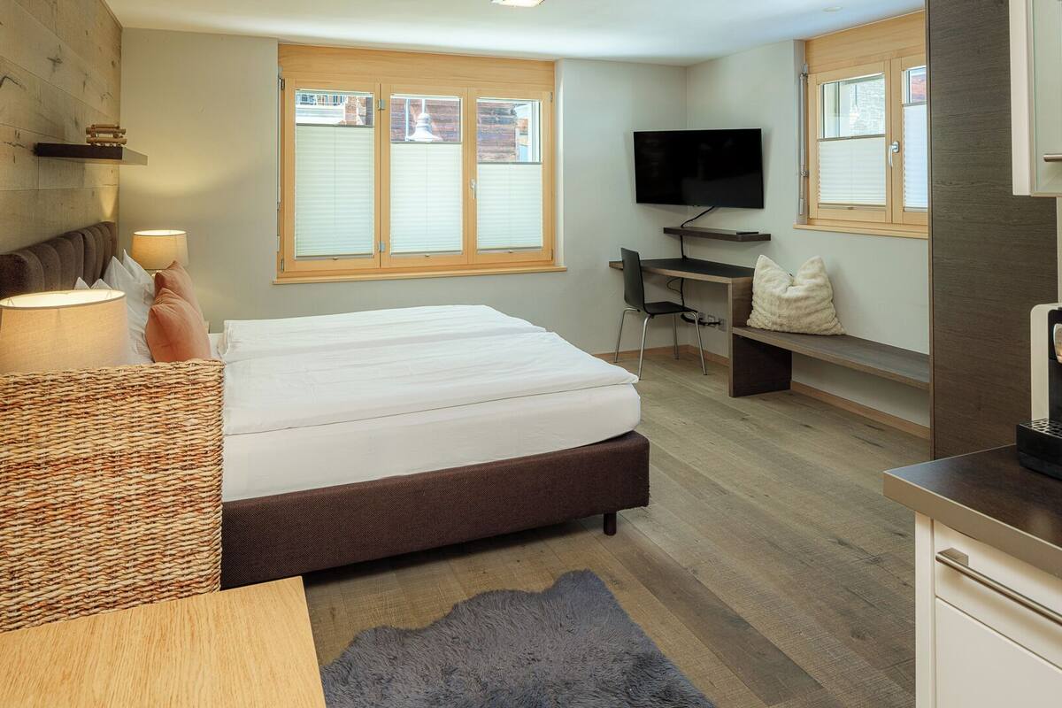 A modern hotel room with a bed, desk, and kitchenette.