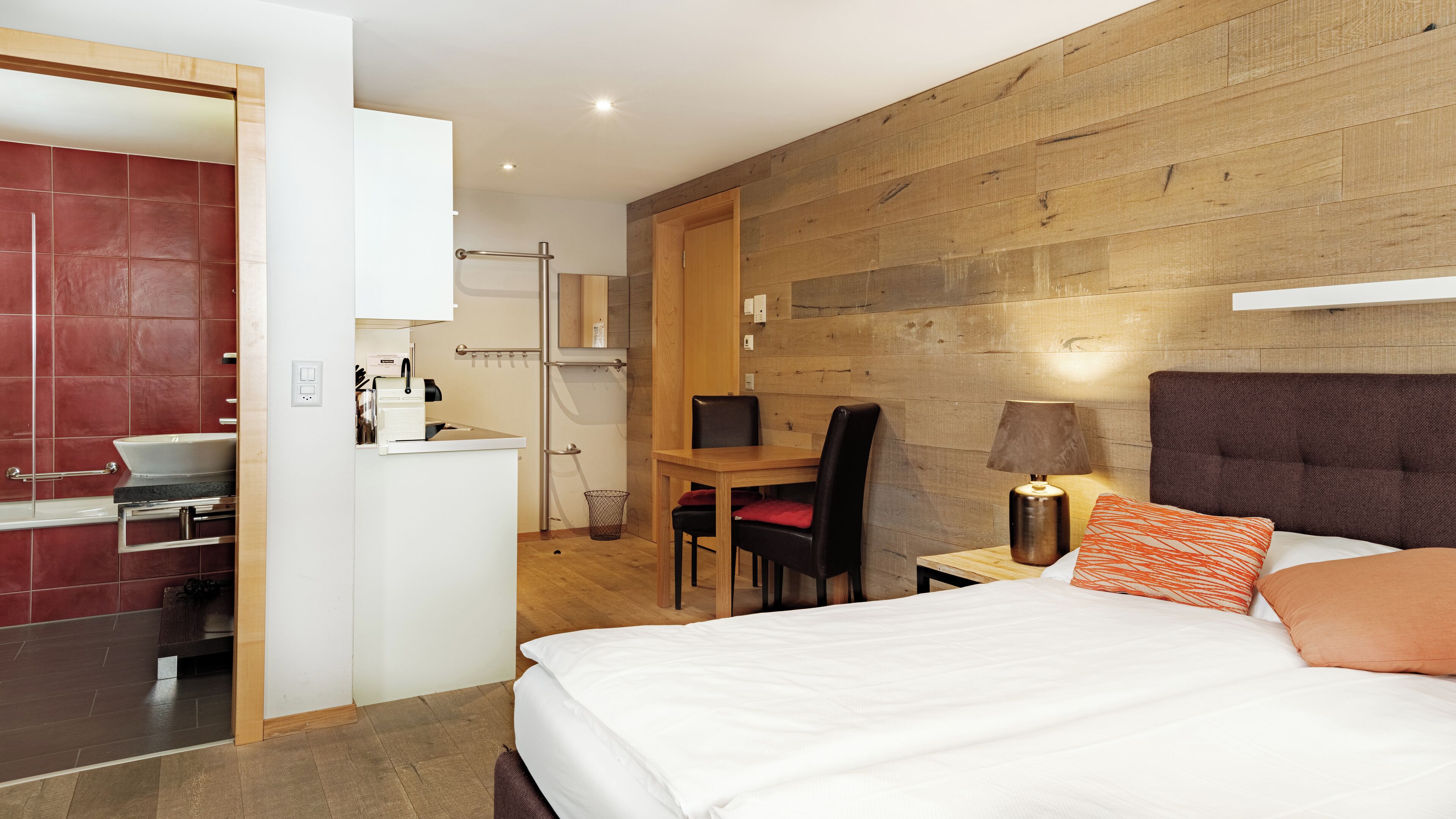 Hotellihuone, jossa on sänky, työpöytä tuolilla, pieni pöytä lampun kanssa ja puinen seinä.