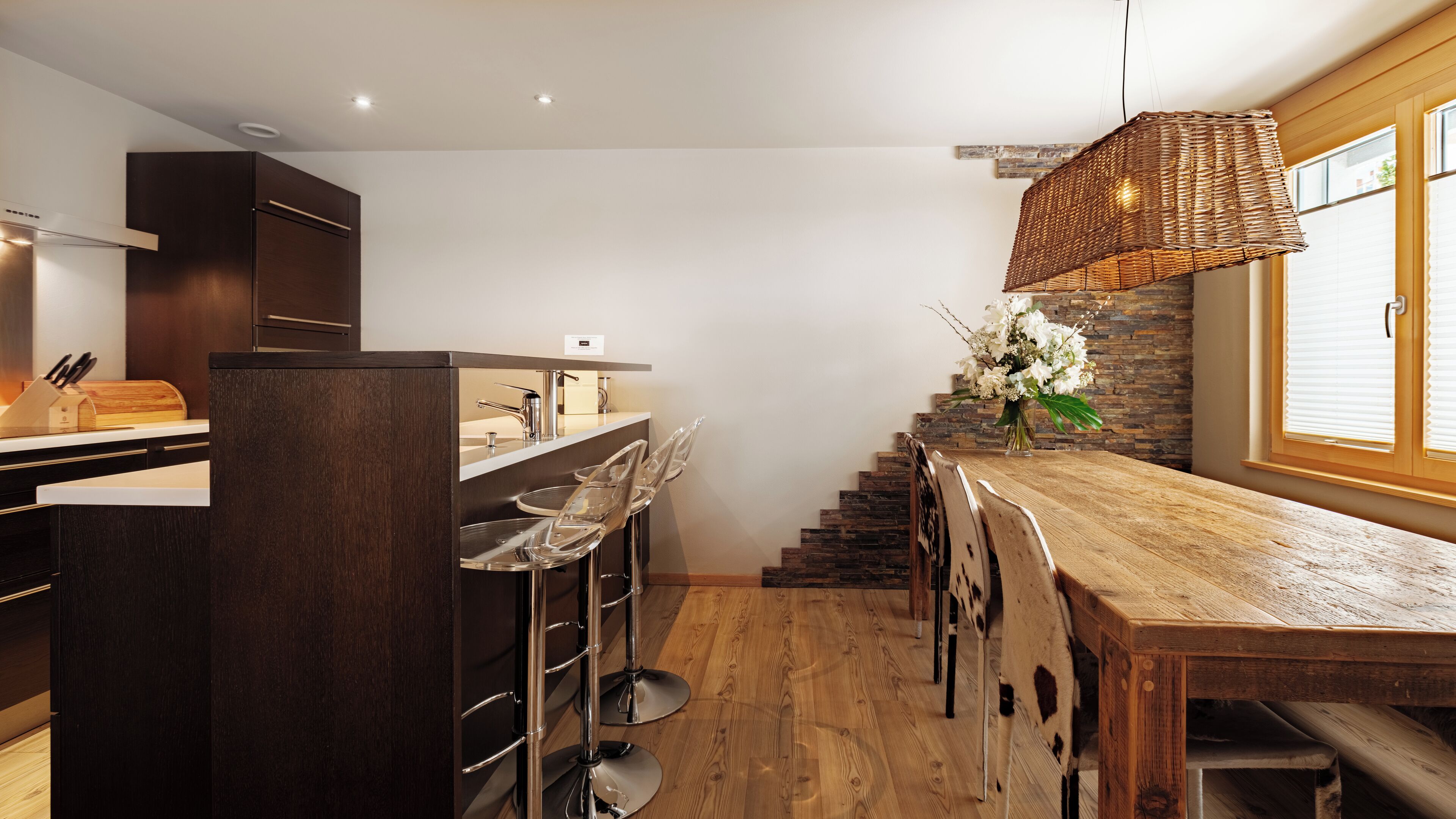 Una cucina moderna con armadietti in legno scuro, un'isola centrale con sgabelli da bar, una lampada a sospensione in vimini e vista all'esterno attraverso una finestra con persiane.
