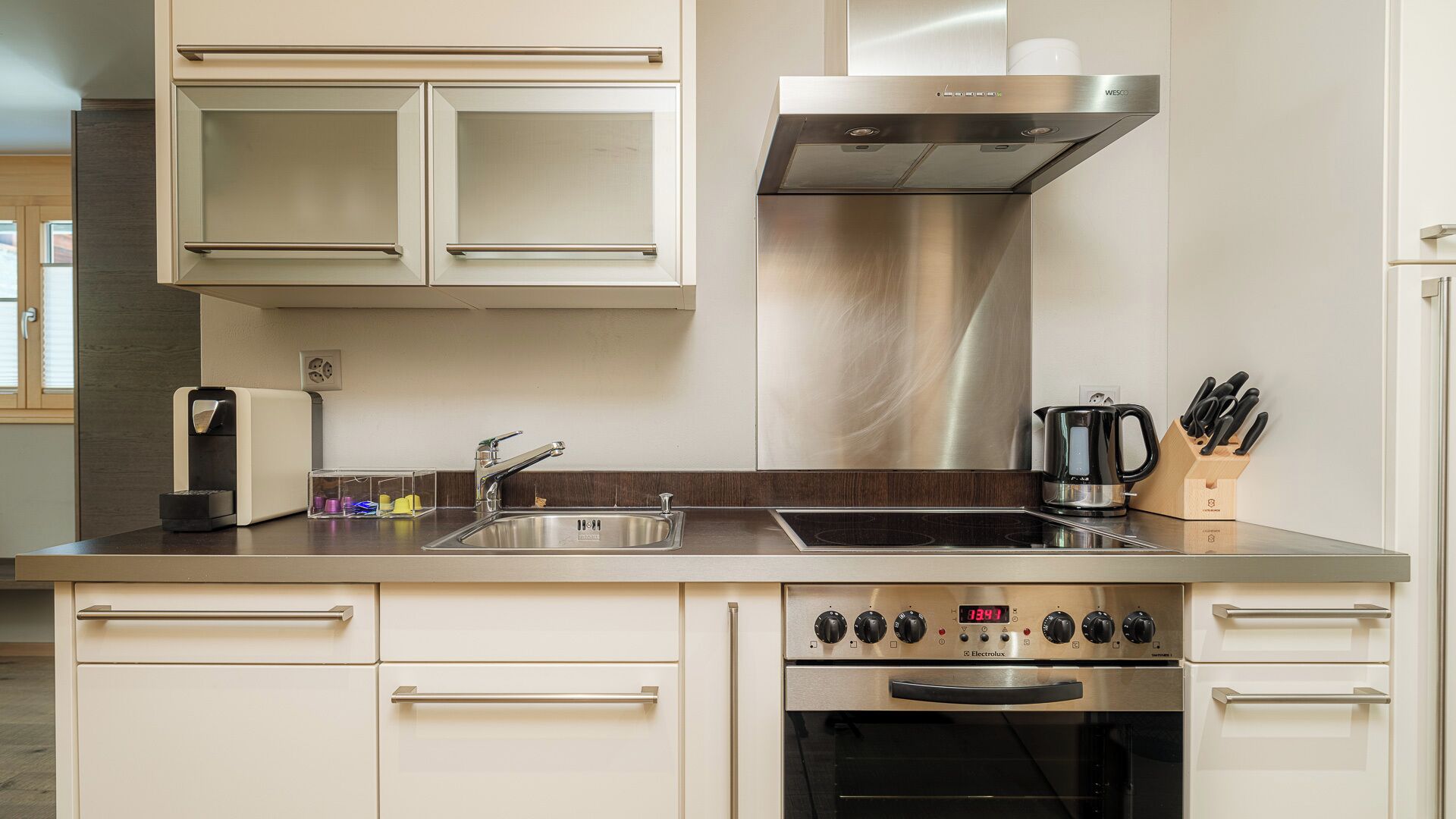 A modern kitchen with a stainless steel range hood, a built-in oven, a double sink, and a coffee maker.