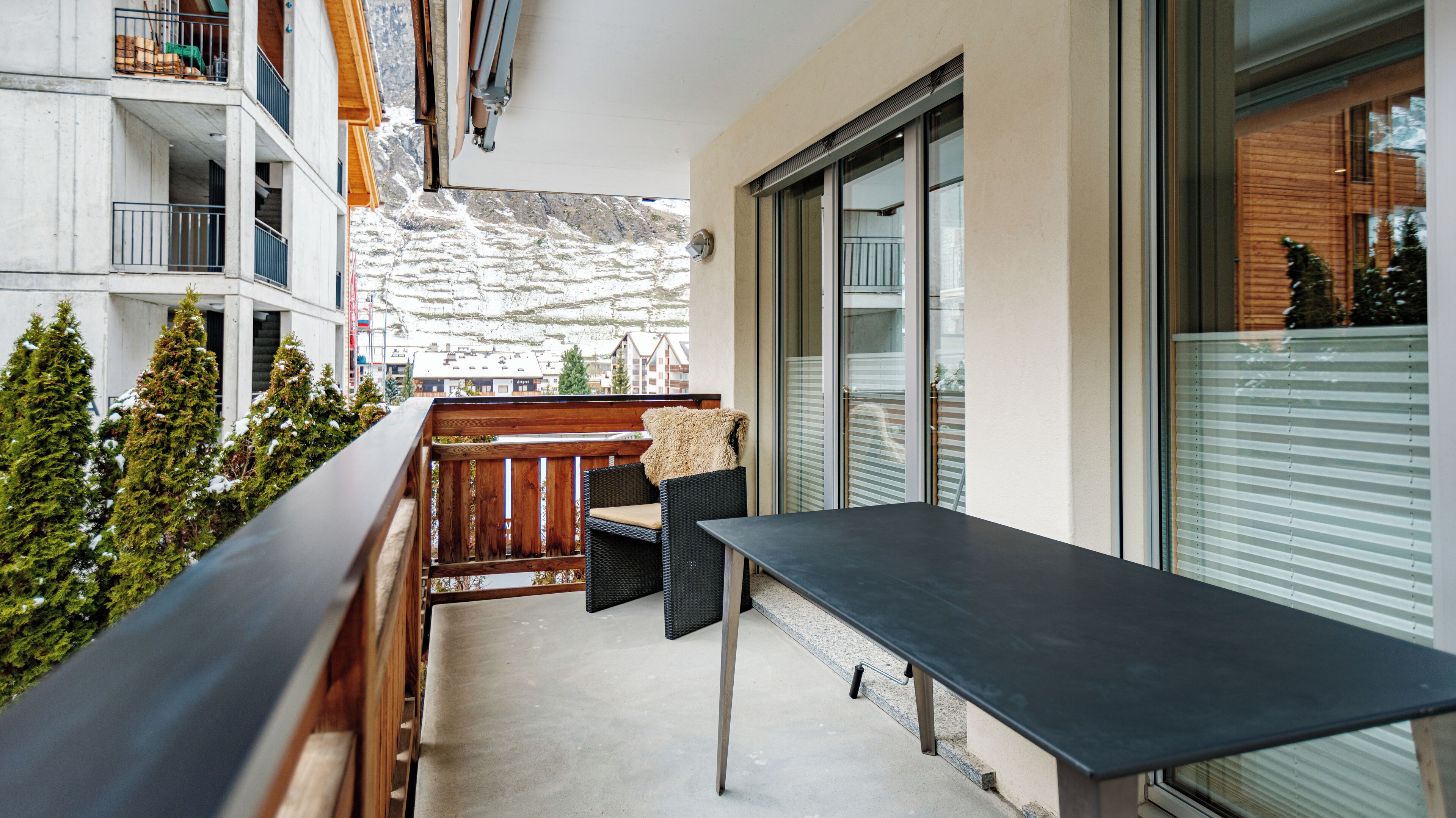 A balcony with a wooden table and chairs, a view of snow-covered mountains, and a building with a glass door.
