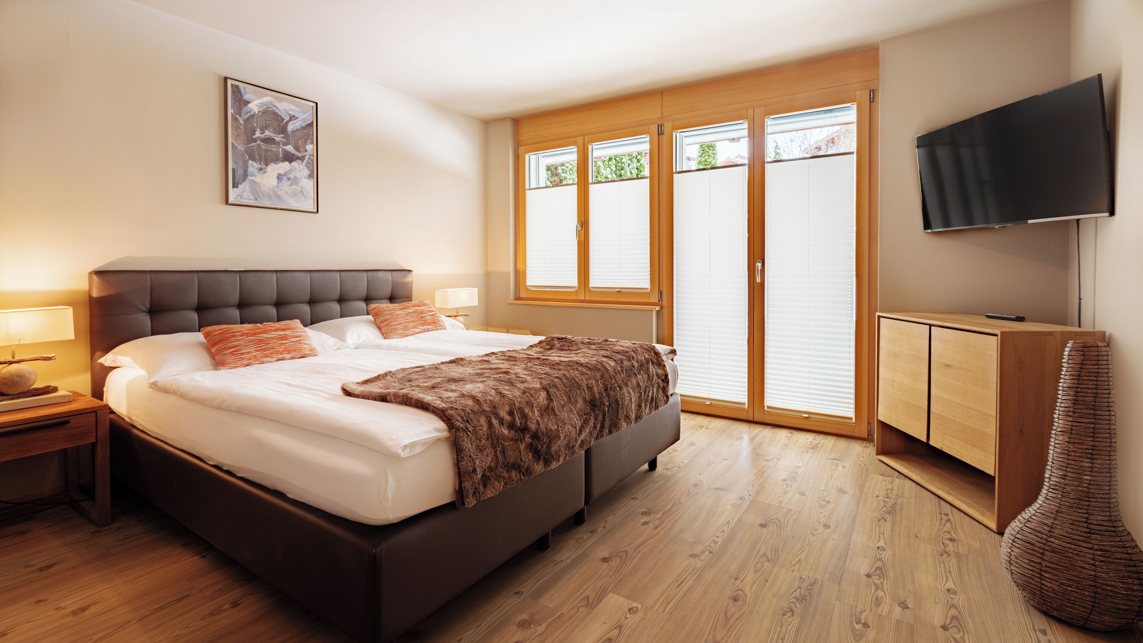A modern bedroom with a large bed, a wooden wardrobe, a television, and a window with blinds.