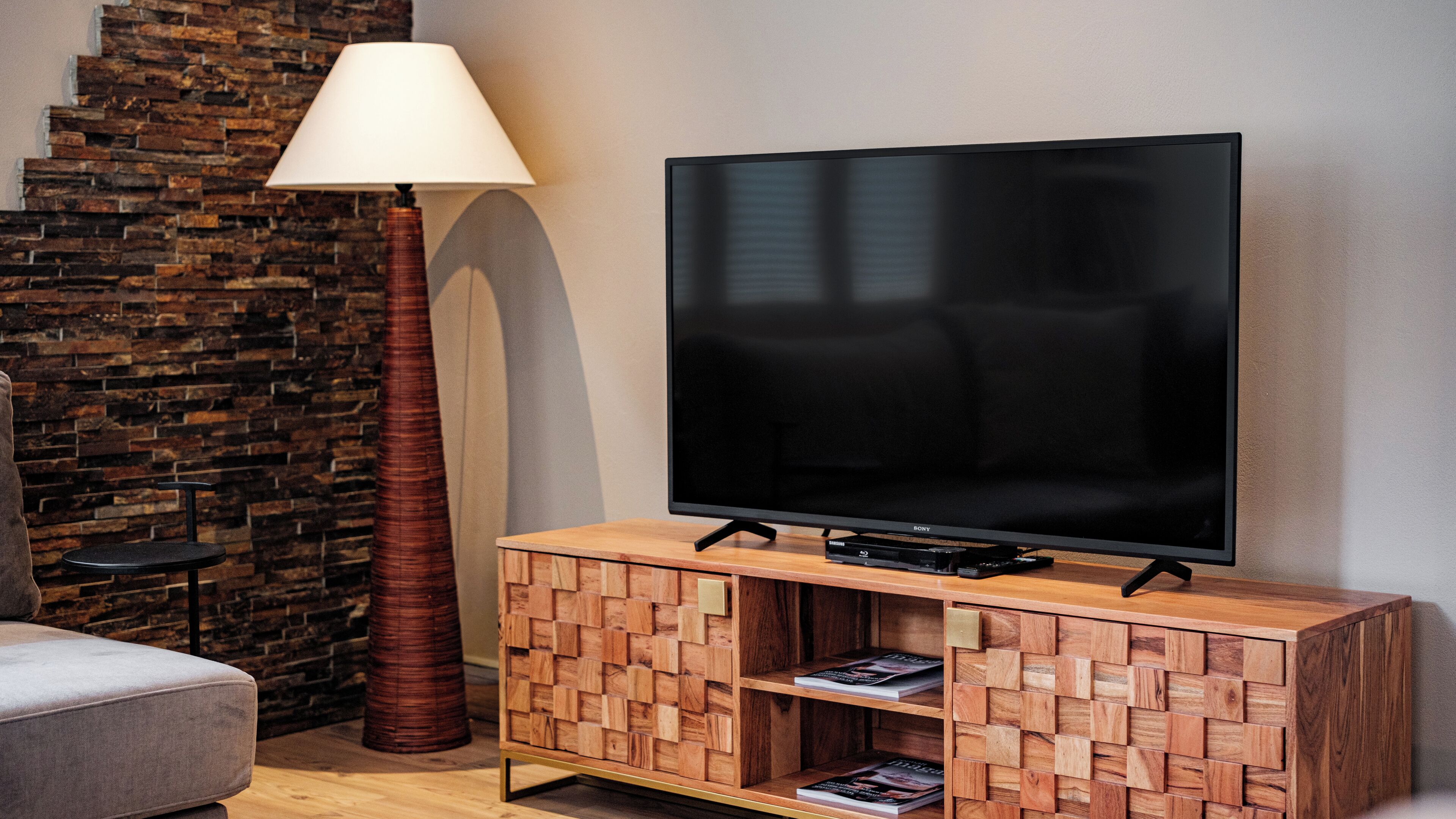 A modern living room with a flat-screen TV on a wooden cabinet, a tall floor lamp, and a stone wall.
