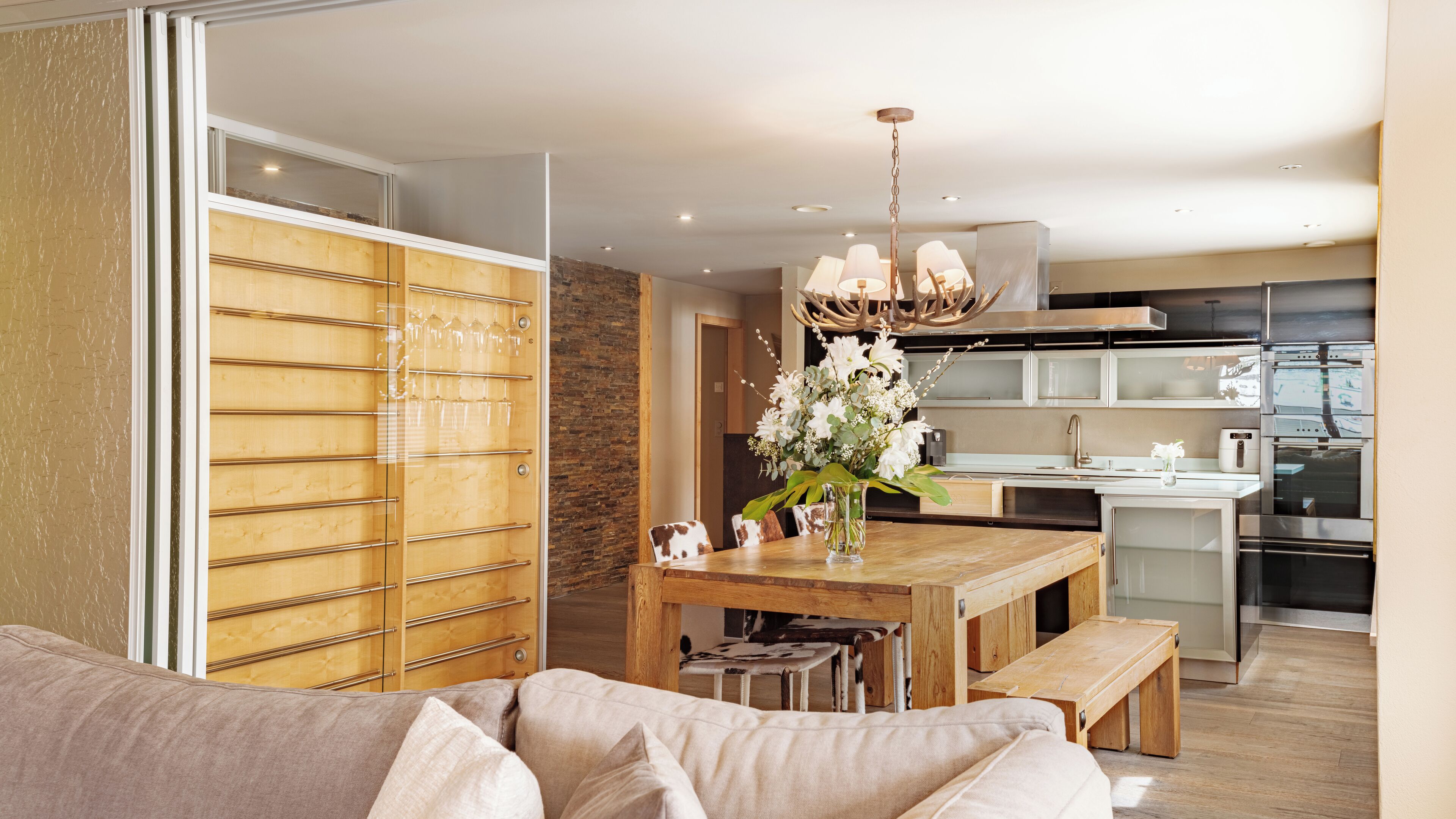 A modern kitchen with stainless steel appliances, a wooden dining table, and a glass-fronted cabinet.