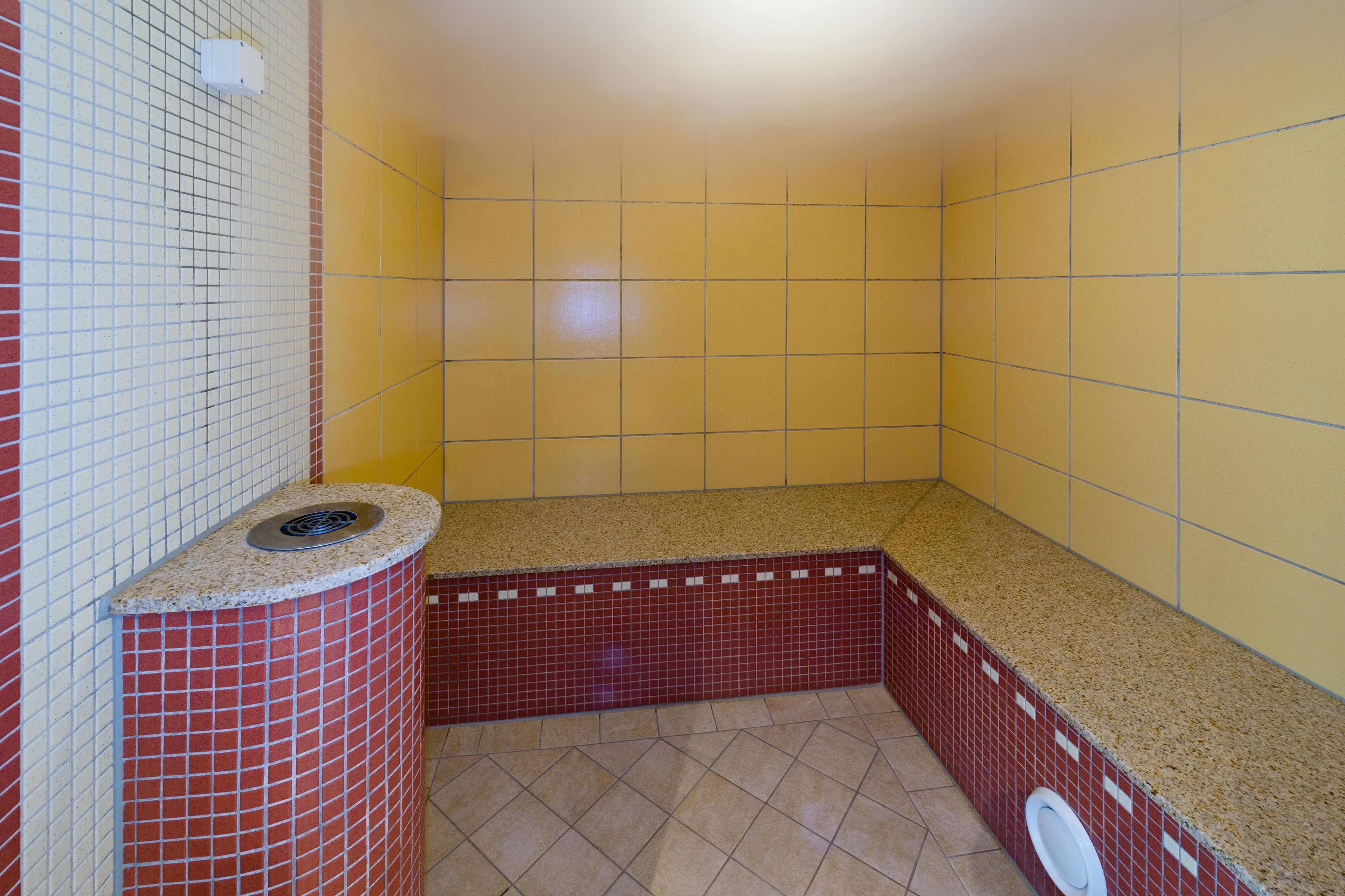 A small, enclosed bathroom with yellow and red tiled walls, a granite countertop, and a visible drain.