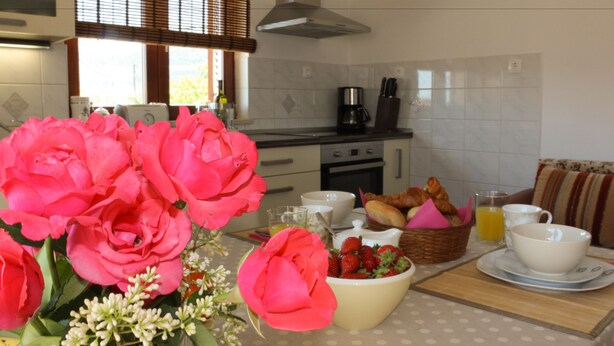 Una cocina con una mesa de comedor lista para el desayuno, con flores, un cuenco de fruta, pan y tazas.